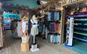 A surf shop featuring beachwear, accessories, and surfboards. A mannequin with a hat and shorts is displayed in the center, surrounded by an assortment of clothing hanging on racks.